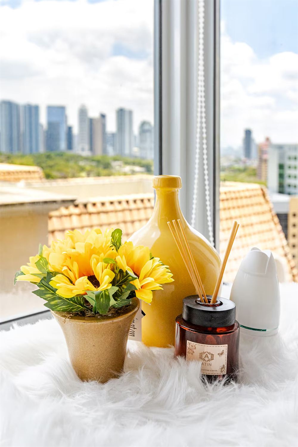A windowsill still life — yellow flowers, a reed diffuser and a glass bottle, with McKinley Hill rooftops and the skyline beyond